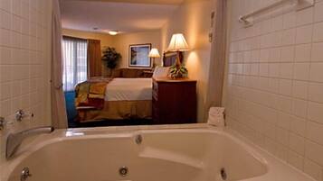 A hotel room with a large bathtub, a bed with a colorful bedspread, a nightstand with a lamp, and a framed picture on the wall.