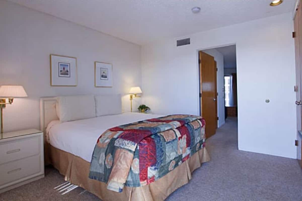 A bedroom with a bed, bedside lamps, a nightstand, and a door leading to another room.