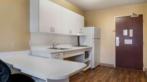 Una pequeña cocina con armarios blancos, un lavamanos, una estufa y un refrigerador. Hay una puerta con cerradura y un interruptor de luz en la pared.