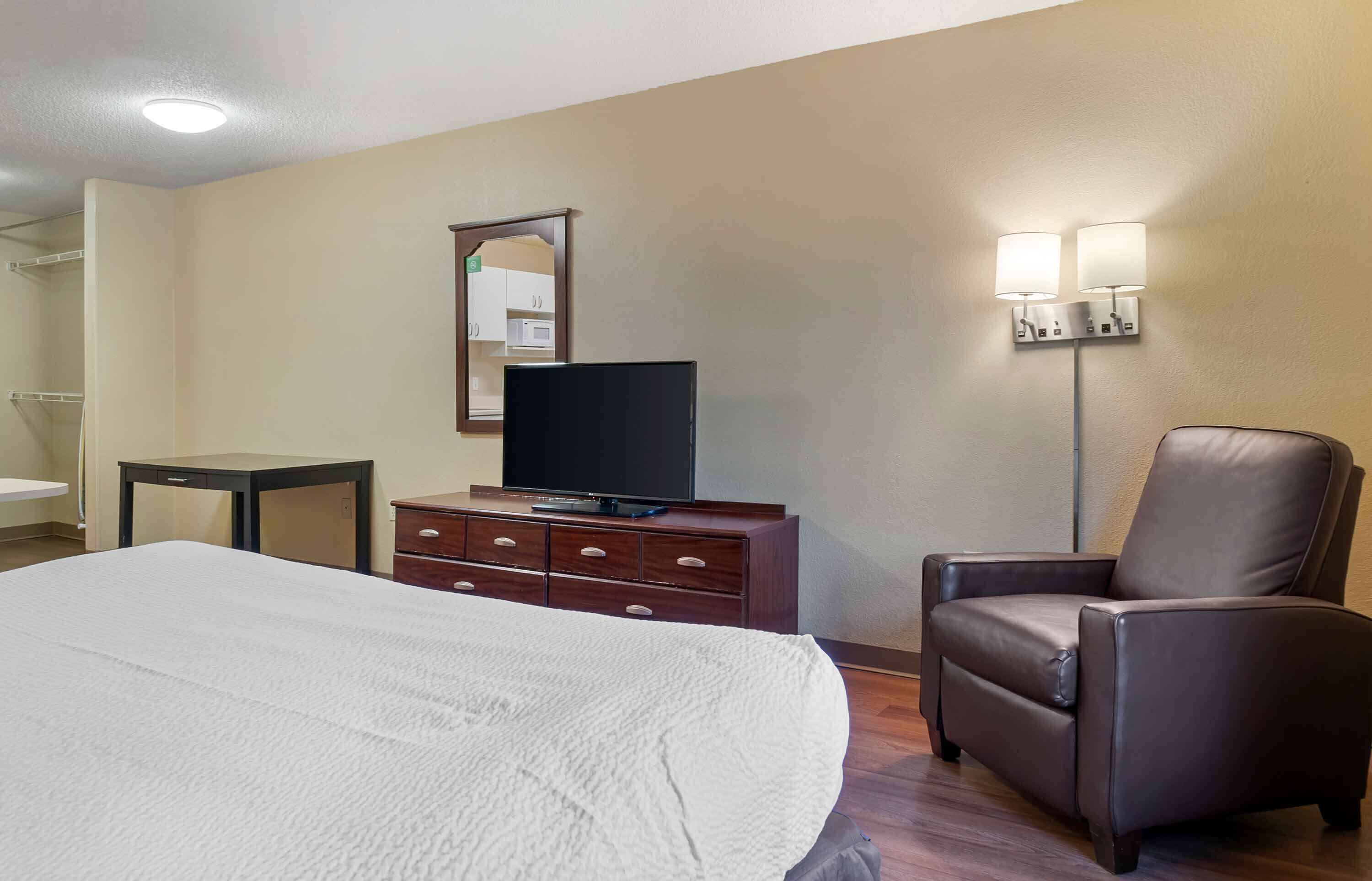 A hotel room with a bed, a dresser with a television, a chair, and a wall-mounted lamp.