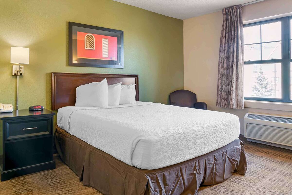 Una habitación de hotel con cama, mesita de noche, lámpara, cuadro enmarco, ventana con cortinas y un equipo de aire acondicionado.