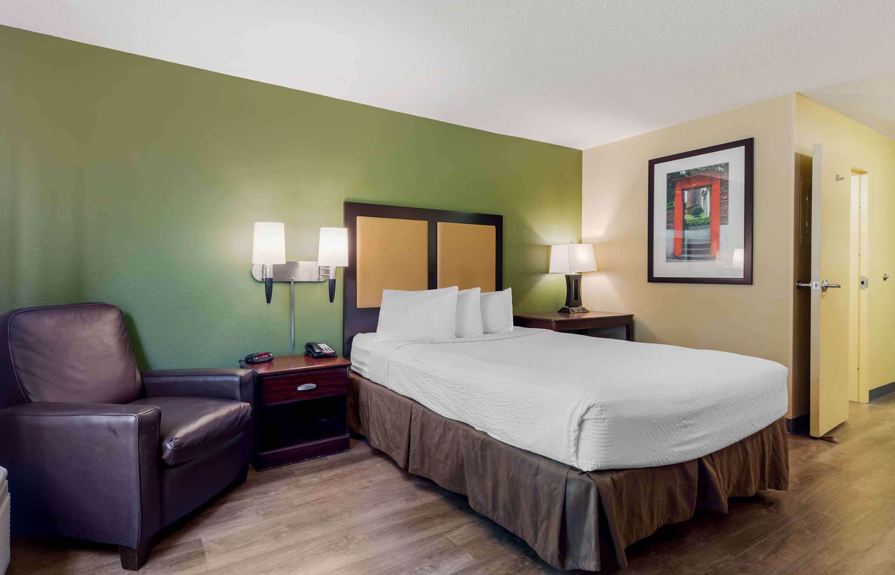 A hotel room with a bed, a chair, bedside tables with lamps, a framed picture on the wall, and a door.