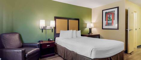 A hotel room with a bed, a chair, bedside tables with lamps, a framed picture on the wall, and a door.