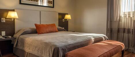 Habitación de hotel ordenada con una cama grande, dos lámparas de noche, un cuadro en la pared y una ventana con cortinas.