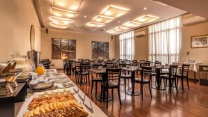 Un comedor con varias mesas y sillas, un bufé con distintas opciones de comida, y amplias ventanas que permiten la entrada de luz natural.