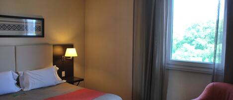 A hotel room with a bed, a nightstand, a lamp, a chair, and a window with curtains.