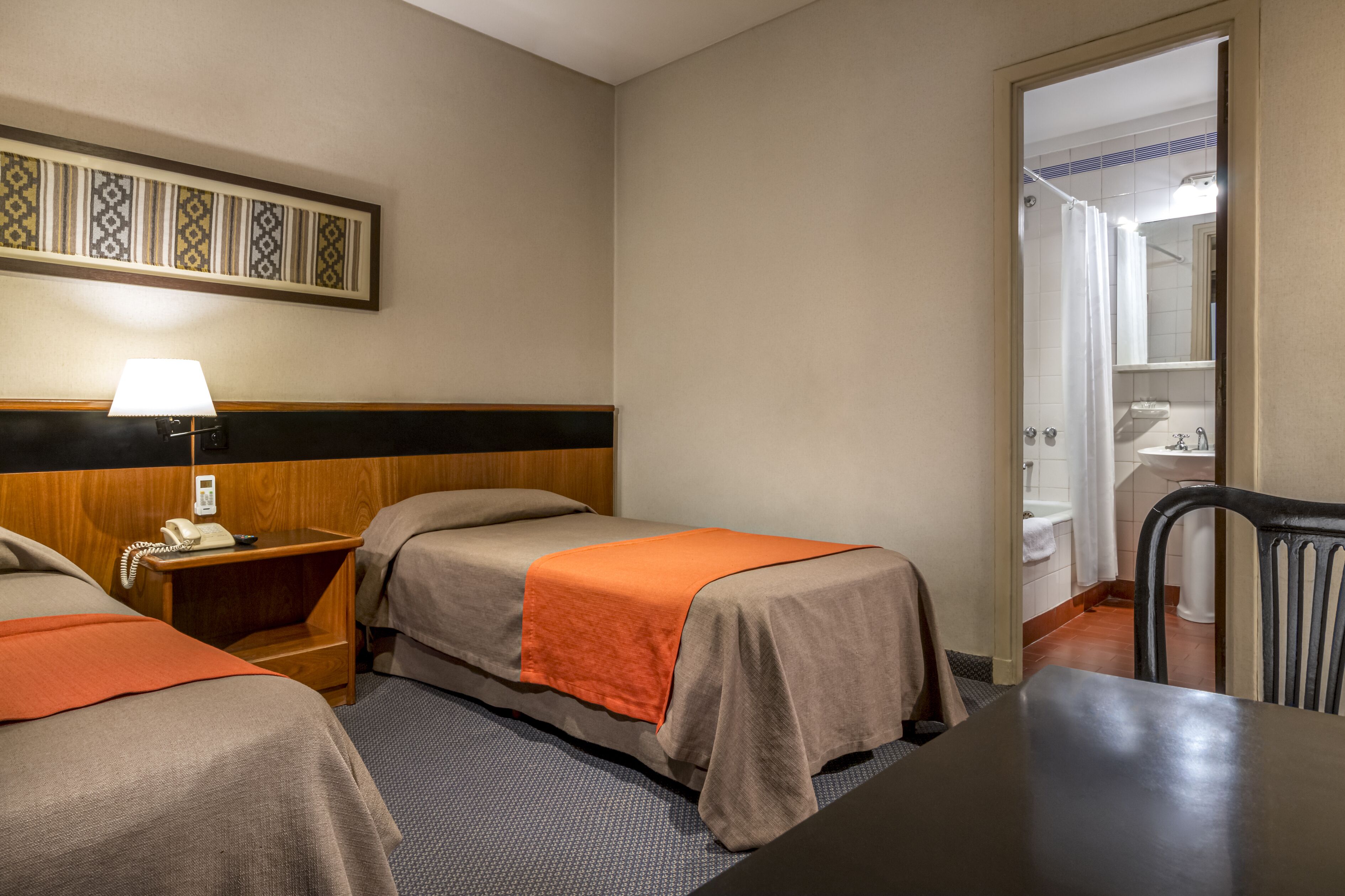 Habitación de hotel con dos camas, un escritorio, una silla, una lámpara y un baño visible a través de una puerta abierta.