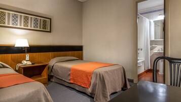 Habitación de hotel con dos camas, un escritorio, una silla, una lámpara y un baño visible a través de una puerta abierta.