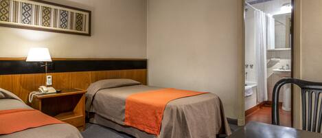 Habitación de hotel con dos camas, un escritorio, una silla, una lámpara y un baño visible a través de una puerta abierta.