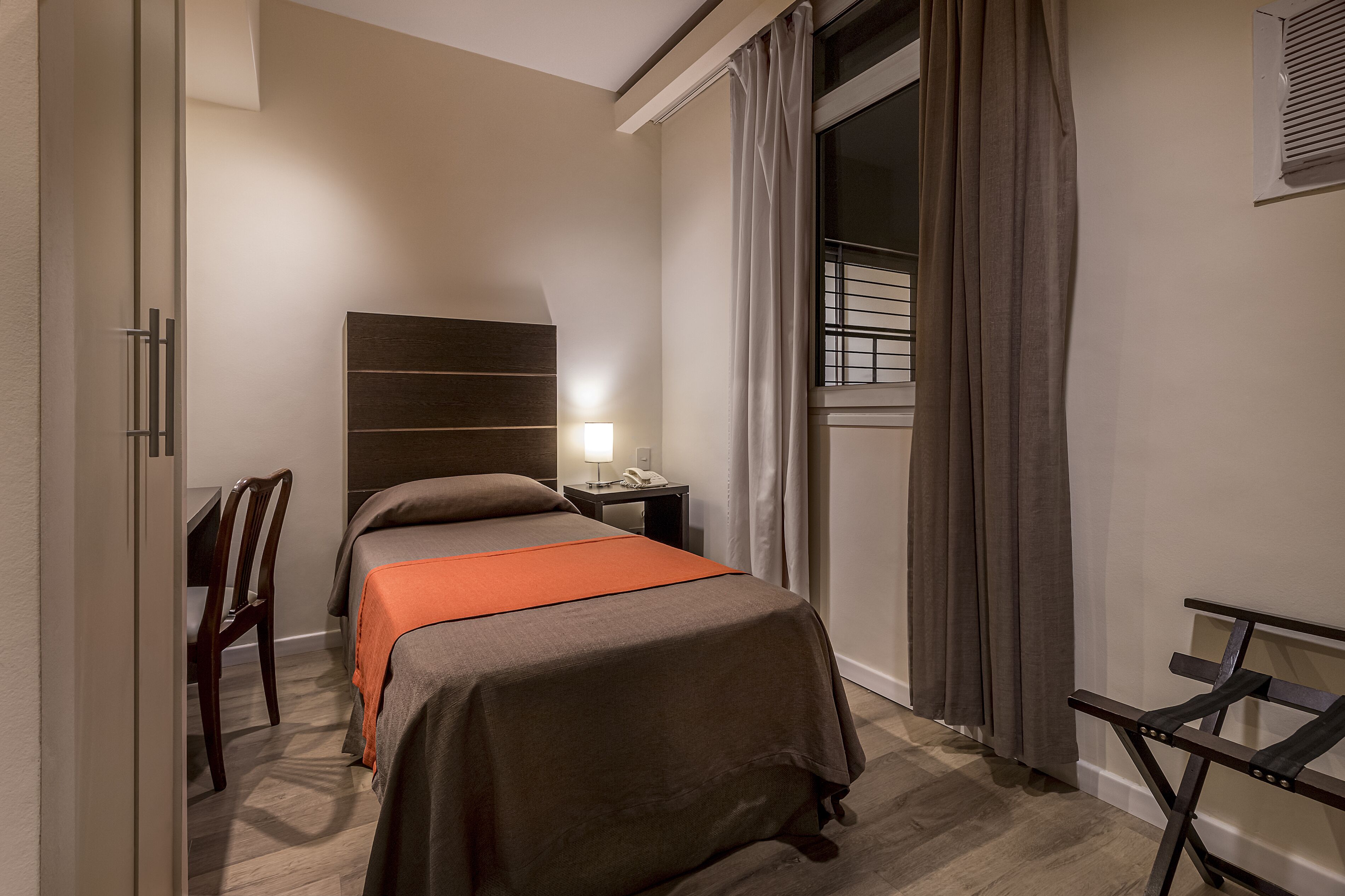 Ein Hotelzimmer mit Bett, Schreibtisch, Stuhl, Nachttisch mit Lampe und einem Fenster mit Vorhängen.