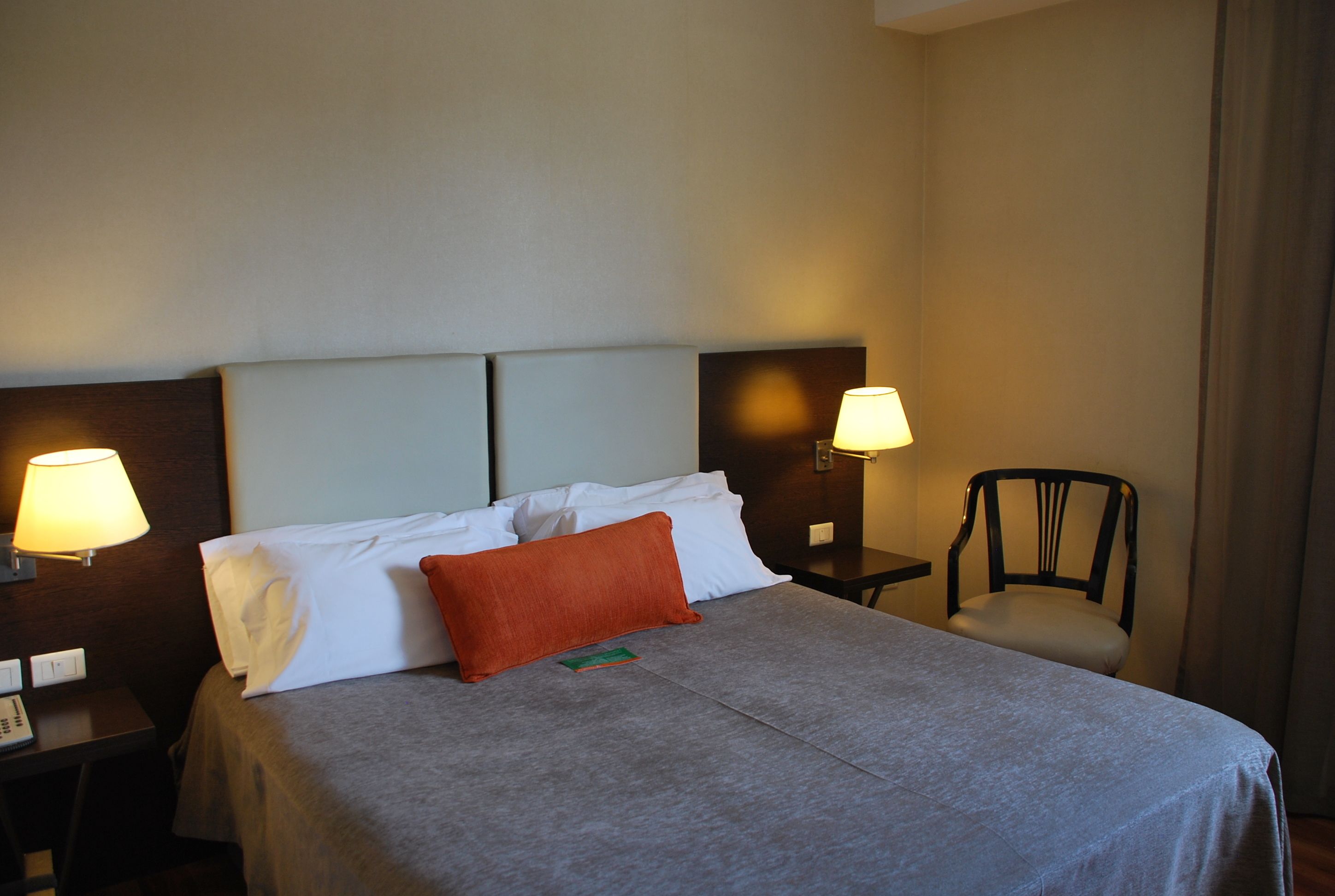 A hotel room with a bed, two bedside lamps, a chair, and a nightstand with a phone.