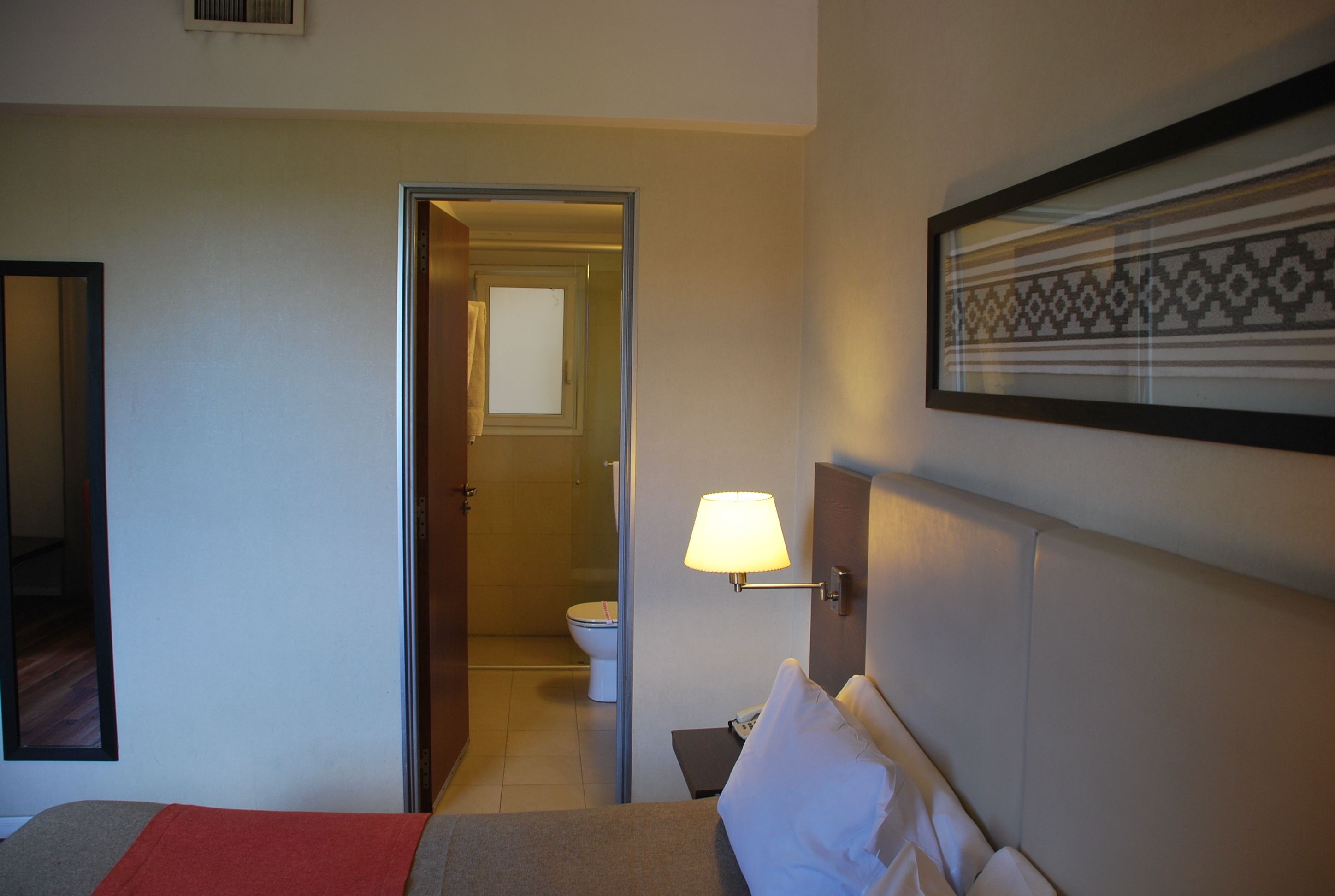 A hotel room with a bed, a nightstand, a wall-mounted lamp, a framed picture, and an open door leading to a bathroom.