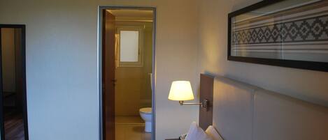 A hotel room with a bed, a nightstand, a wall-mounted lamp, a framed picture, and an open door leading to a bathroom.
