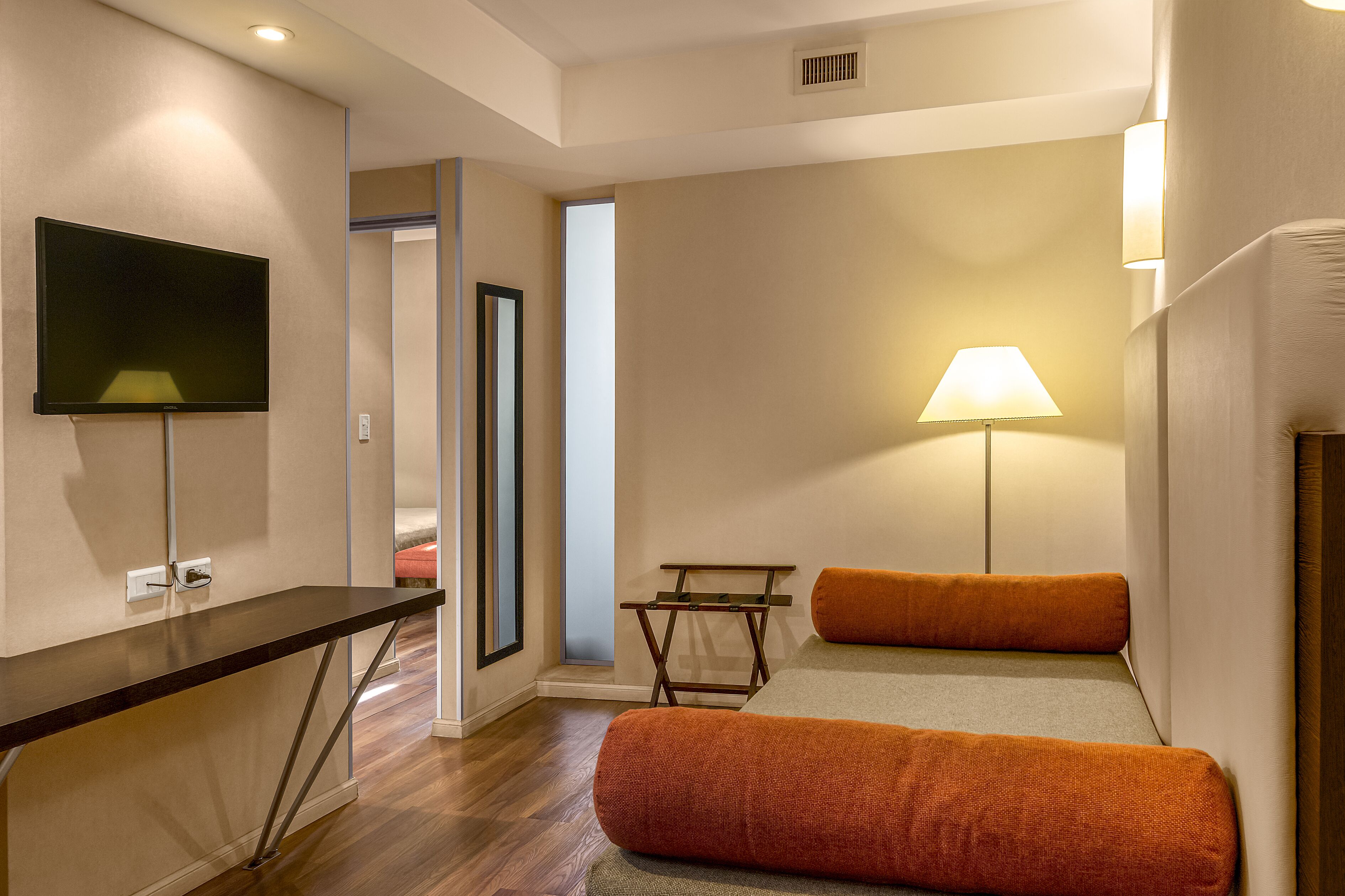 Habitación de hotel moderna con televisor de pantalla plana, una mesita, una lámpara y una cama con almohadas anaranjadas.