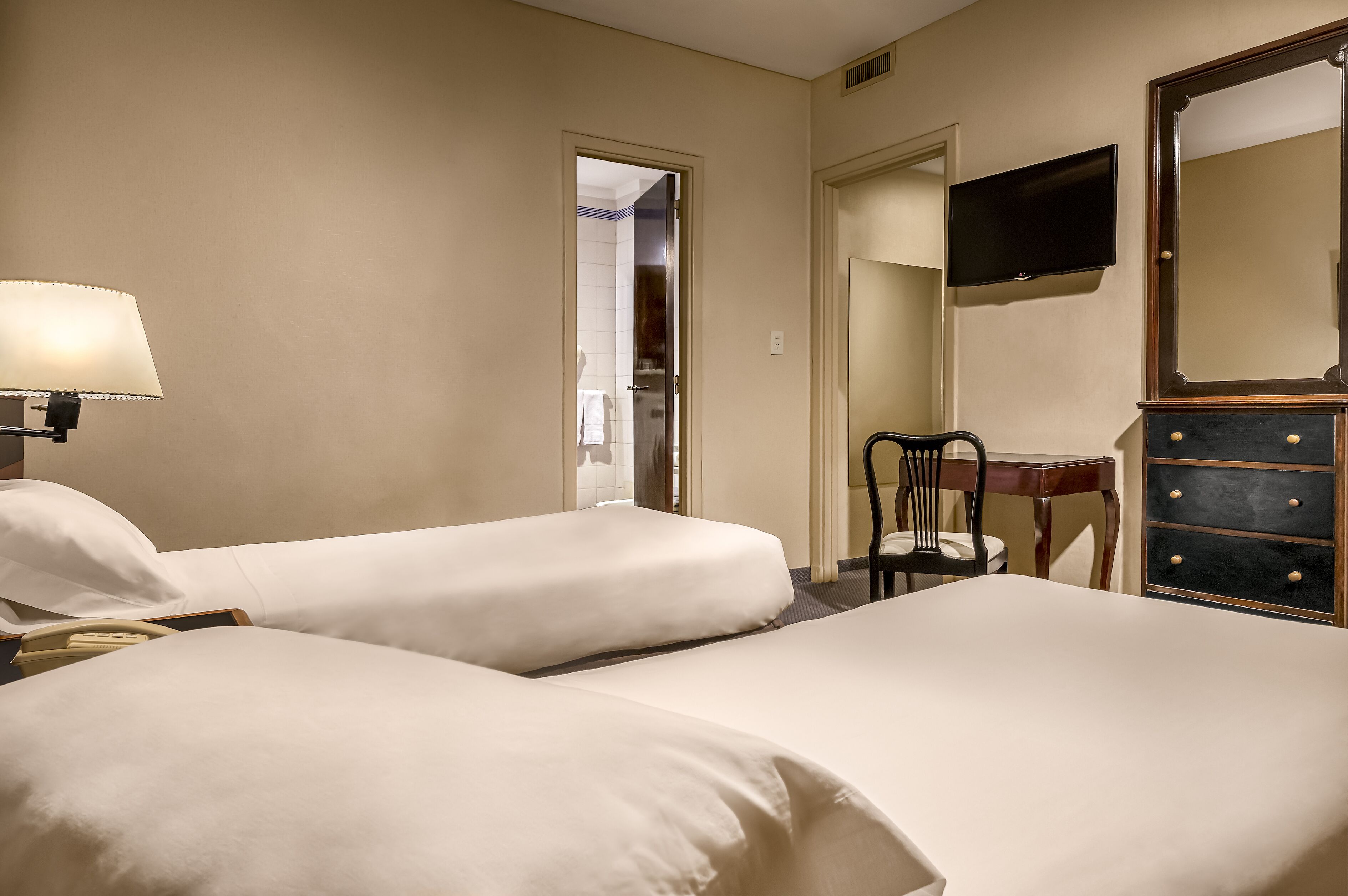 A hotel room with two beds, a desk, a chair, a dresser, and a TV on the wall.