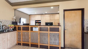 A hotel reception area with a wooden counter, a coffee machine, and a sign indicating a 24-hour check-in service. - Howard Johnson by Wyndham National City/San Diego South (National City)