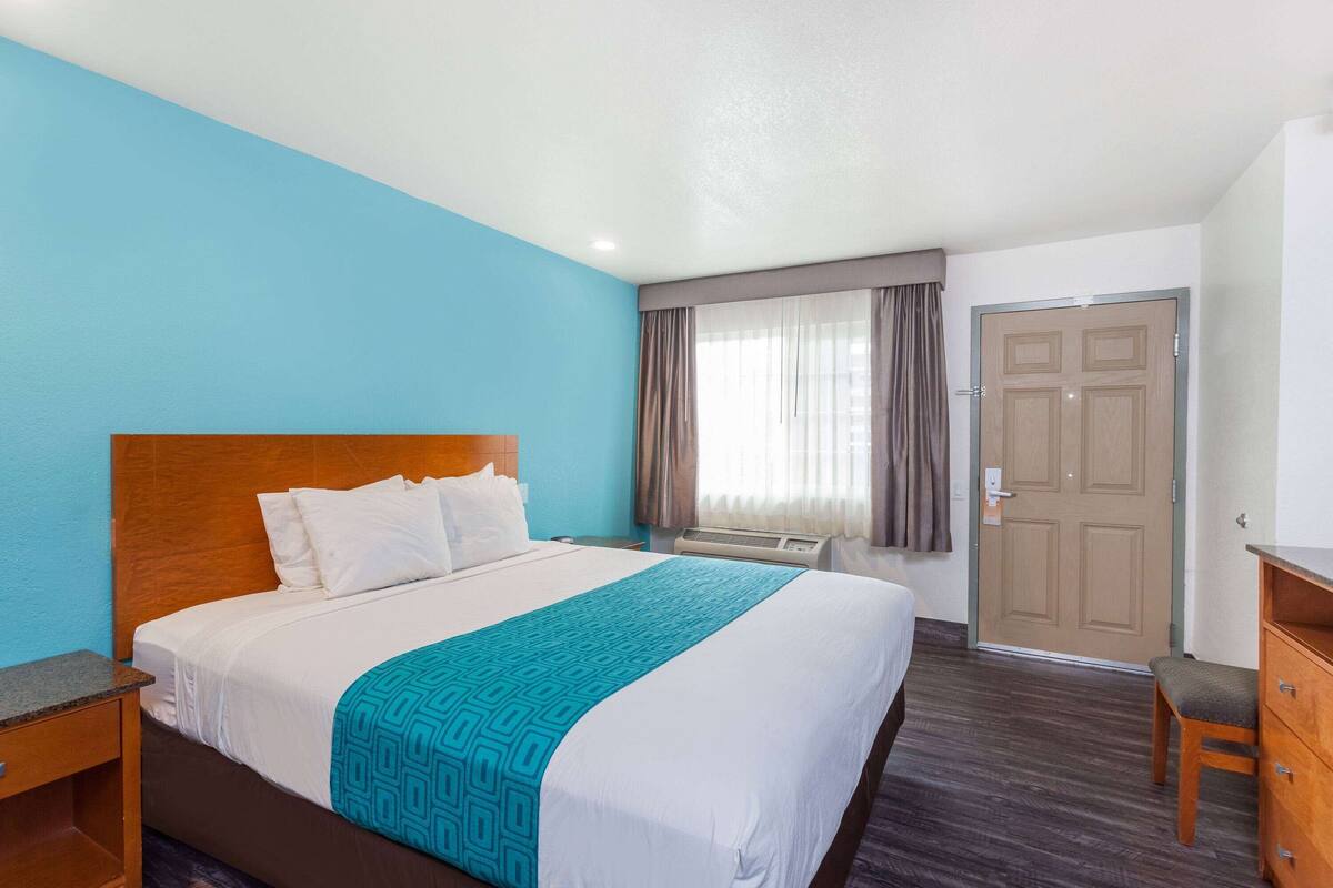 Une chambre d’hôtel avec un grand lit, une tête de lit en bois, un mur d’accent bleu, une fenêtre avec des rideaux, une porte et une petite table de chevet en bois.
