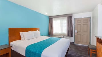 Habitación de hotel con una cama grande, cabecera de madera, pared azul de acento, ventana con cortinas, una puerta y una mesita de noche de madera.