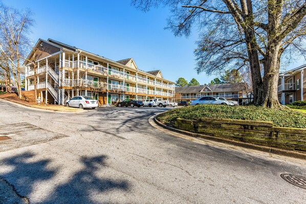 A multi-story hotel with balconies, a parking lot, and a tree-lined street. - HomeTowne Studios by Red Roof Atlanta - Norcross Downtown (Norcross)