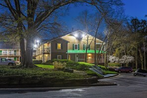 A two-story building with a green roof and multiple windows, surrounded by trees and parked cars.