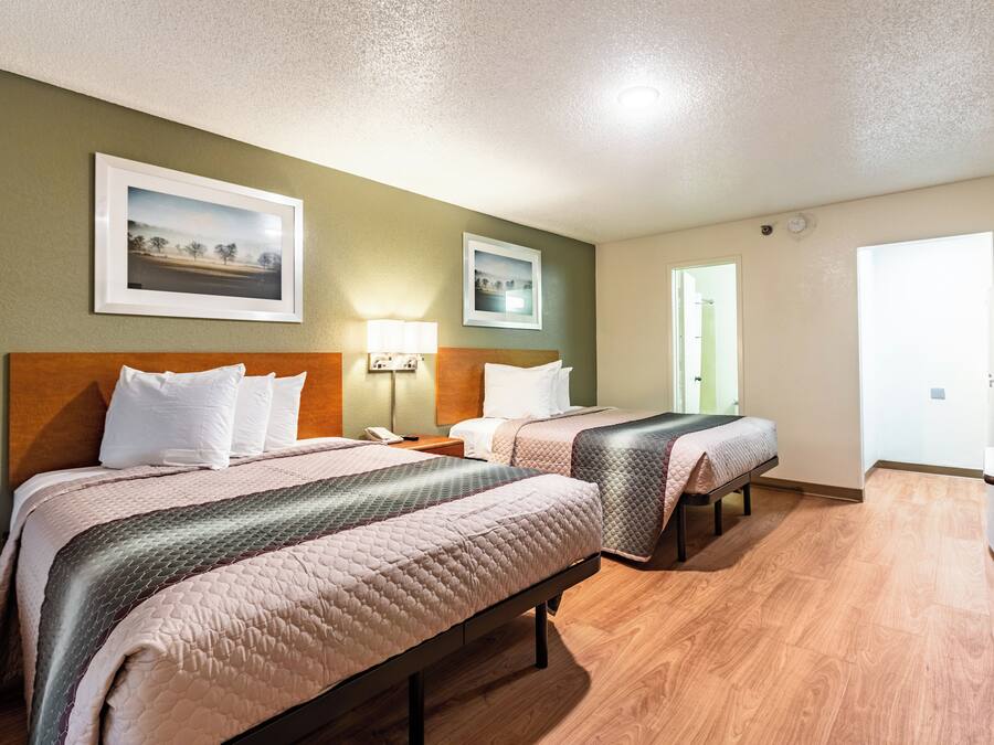 Habitación de hotel con dos camas, un escritorio, una silla, una lámpara y dos cuadros en la pared.