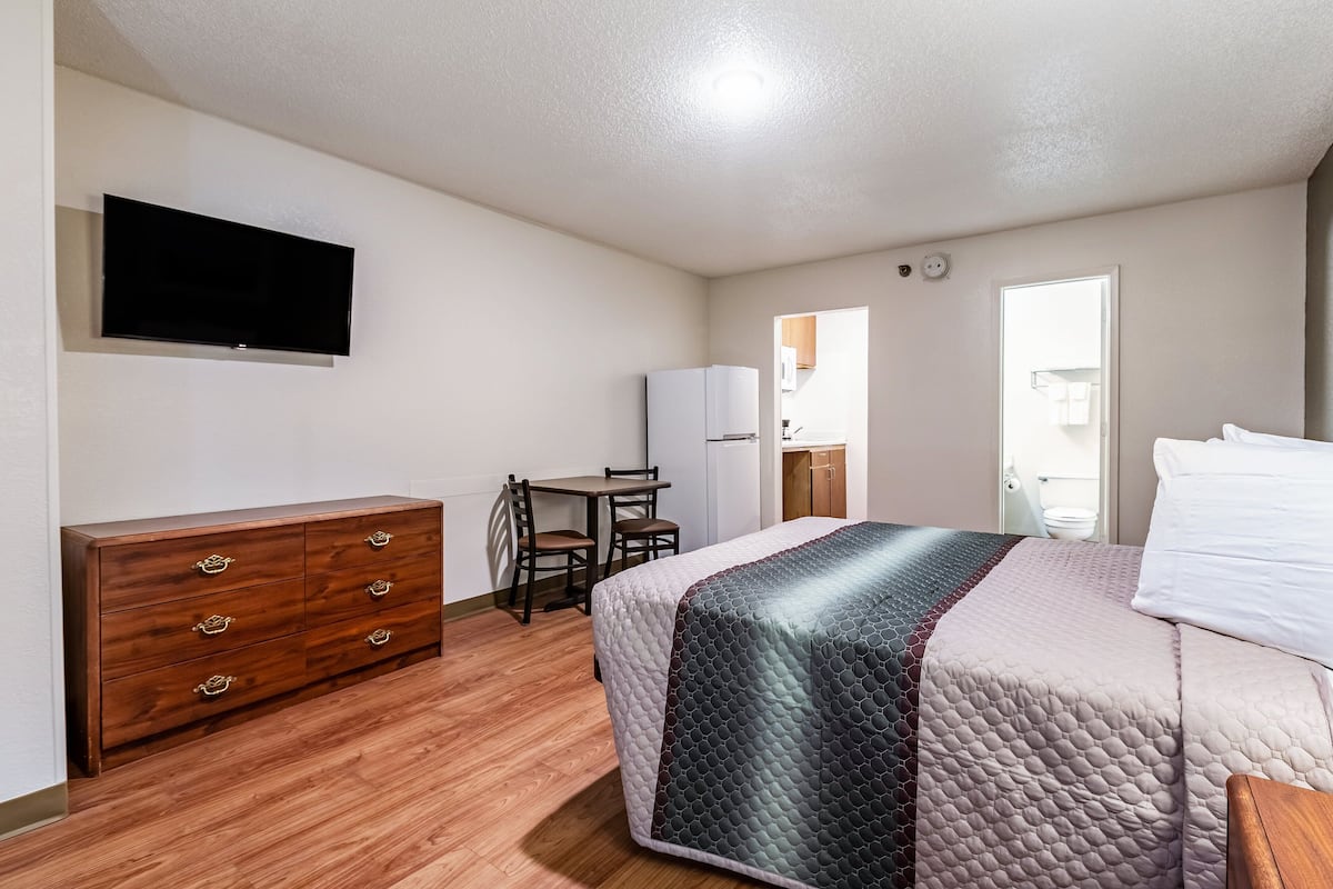 A bedroom with a bed, a wooden dresser, a television mounted on the wall, a small table with chairs, and a door leading to a bathroom.