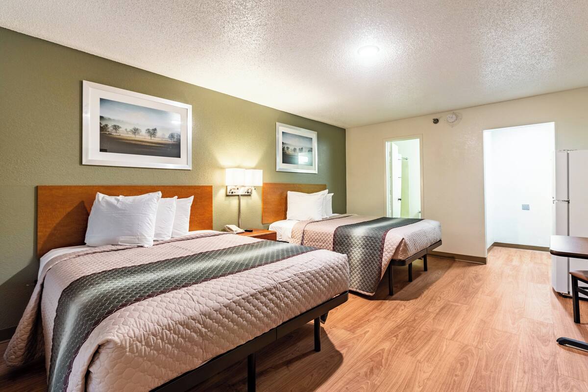 A hotel room with two beds, a desk, and a chair. There are framed pictures on the wall, a lamp, and a phone on the bedside table.