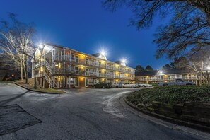 A multi-story hotel with outdoor lighting, a parking area, and a landscaped garden. - HomeTowne Studios by Red Roof Atlanta - Norcross Downtown (Norcross)