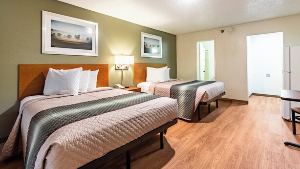 A hotel room with two beds, a desk, and a chair. There are framed pictures on the wall and a door leading to another room. - HomeTowne Studios by Red Roof Atlanta - Norcross Downtown (Norcross)