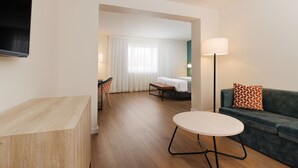 Una habitación de hotel moderna con un escritorio de madera, una mesa redonda, un sofá con cojines estampados, una cama con ropa de cama blanca y un ventanal con cortinas traslúcidas.
