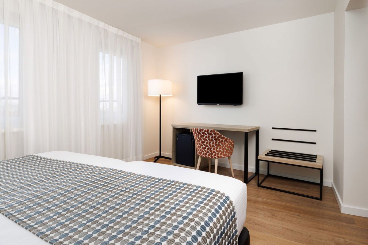 A hotel room with a bed, a desk, a chair, a TV, and a lamp.