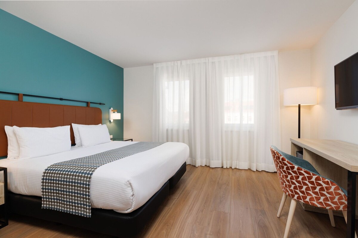 Ett hotellrum med en säng, ett skrivbord, en stol, en tv och ett fönster med gardiner.