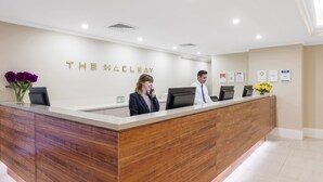 Un comptoir d’accueil avec un revêtement en bois et un plan de travail blanc. Le mur derrière porte l’inscription « THE MAELAY » en lettres dorées.