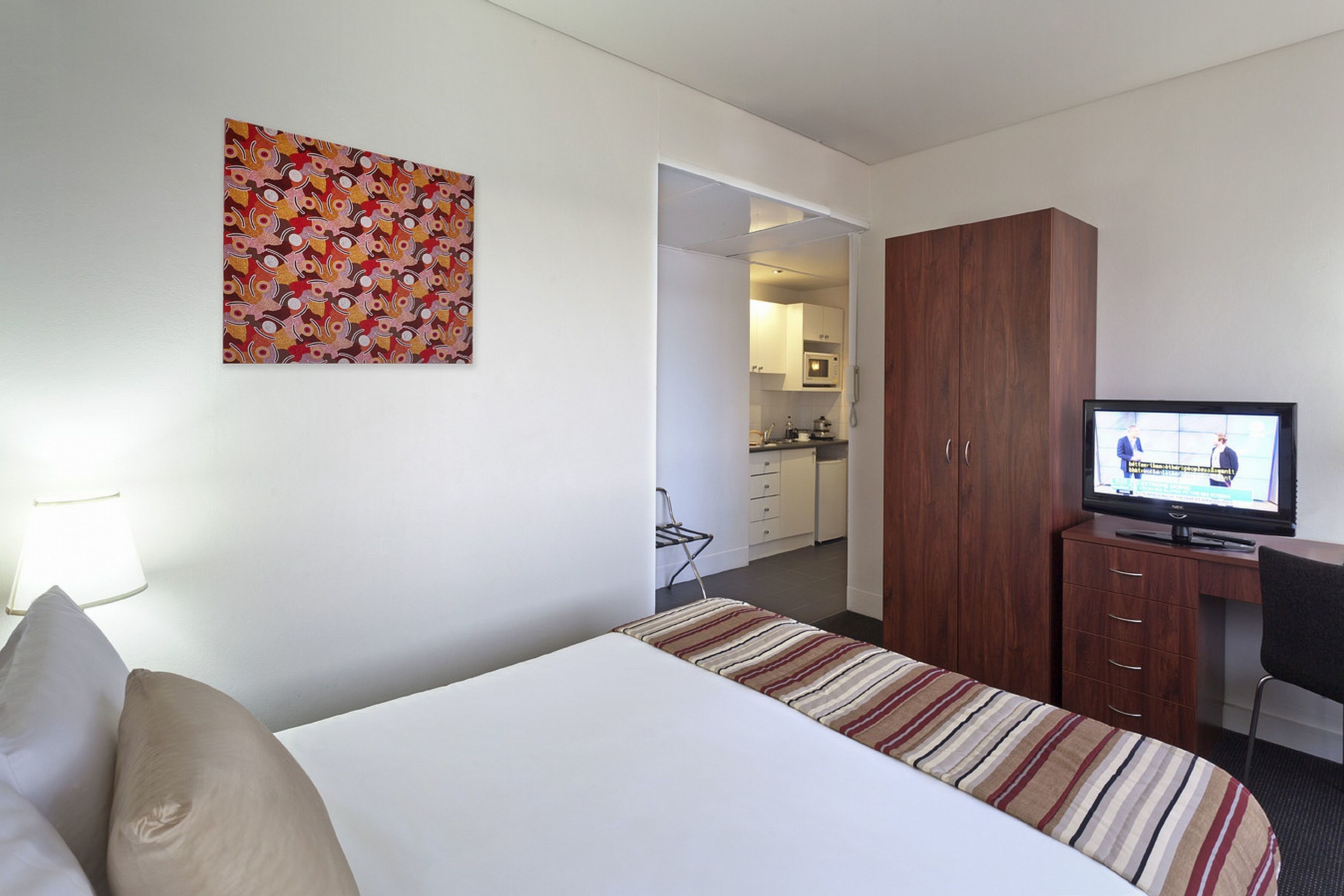 A hotel room with a bed, a television, a desk, and a kitchen area visible through an open door.