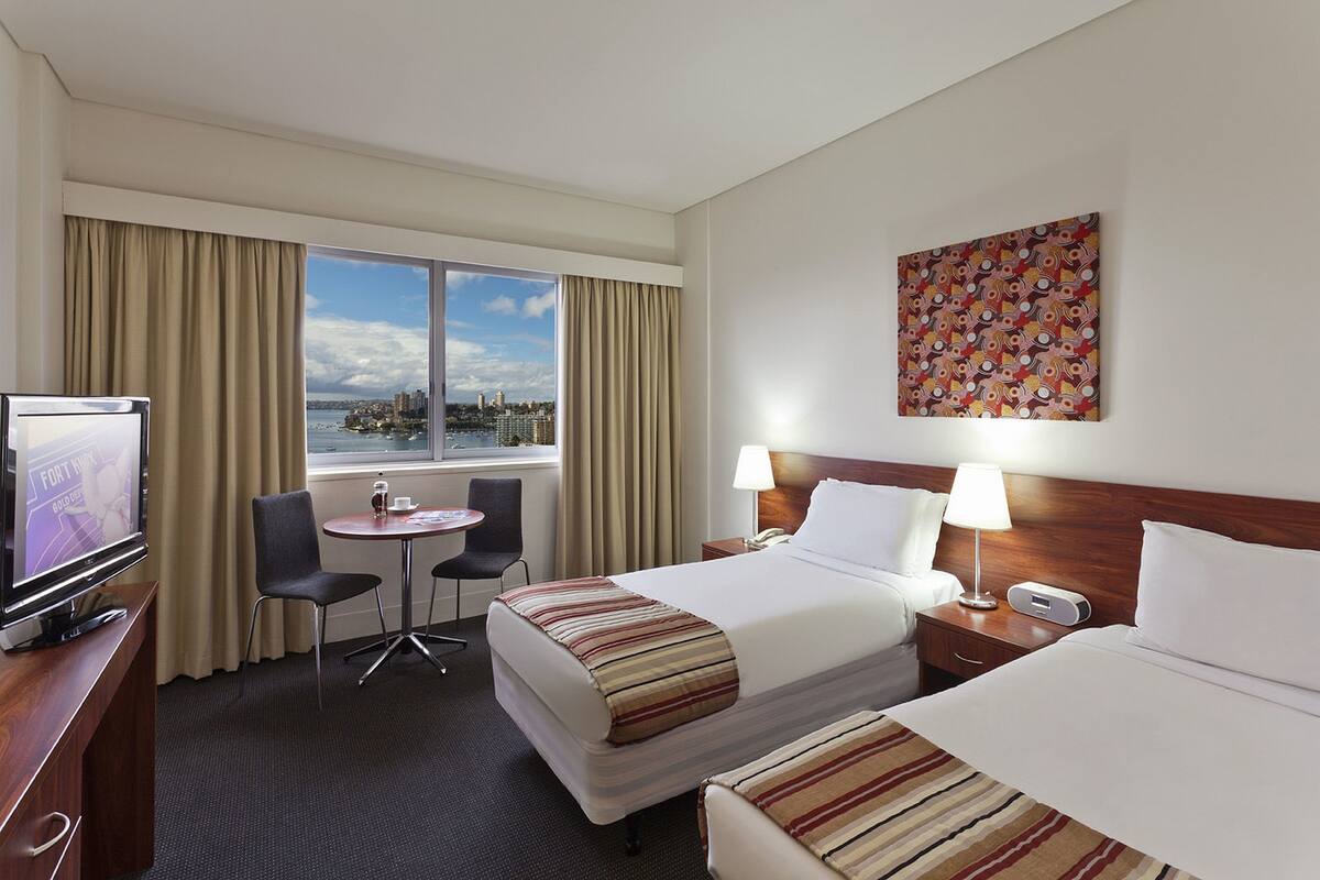 Habitación de hotel con cama, escritorio con televisor, una silla, una mesita y ventana con cortinas, ofreciendo vista a la ciudad.