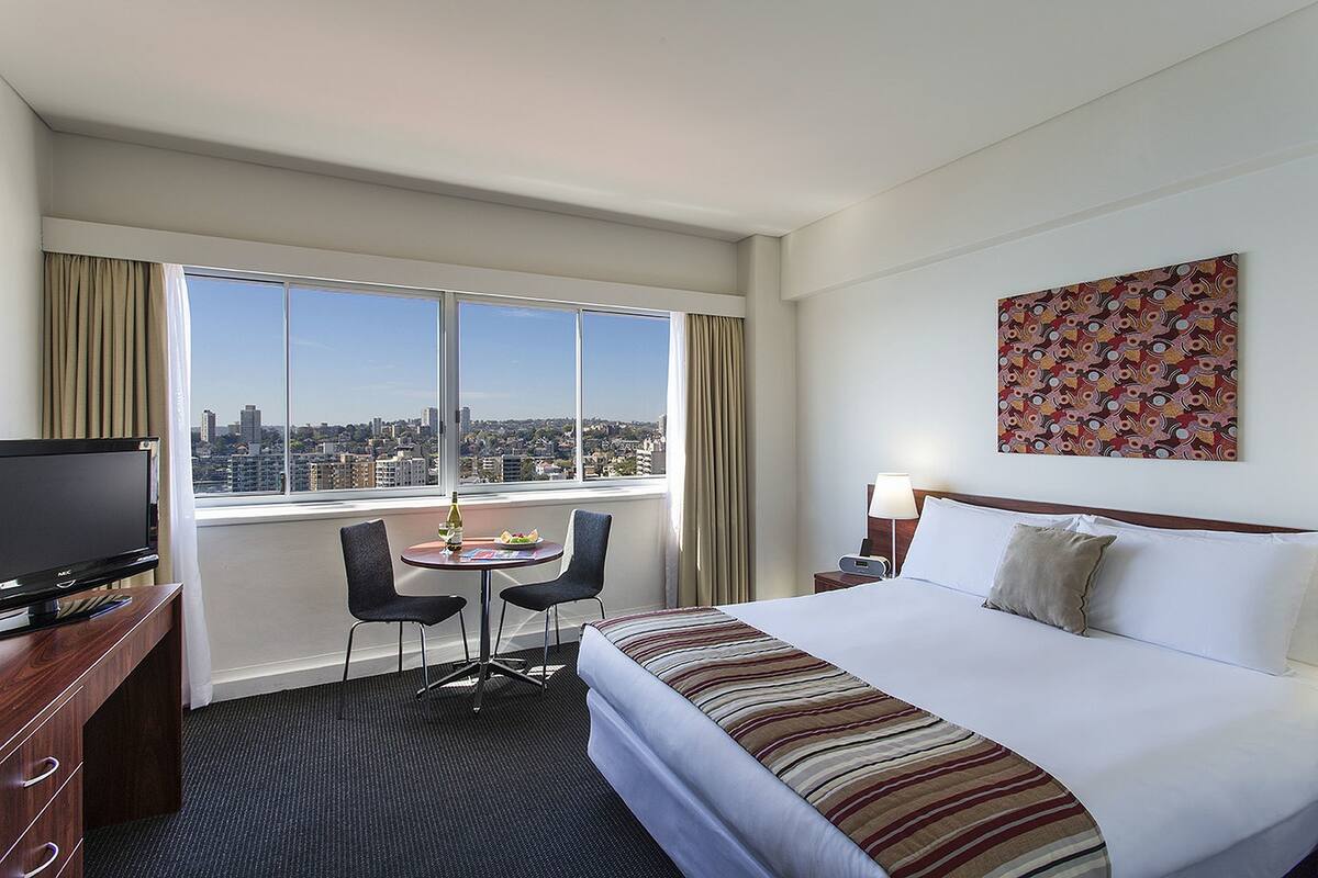Habitación de hotel con un ventanal grande, una cama, un escritorio con televisión, una silla y una mesa con botella y fruta.