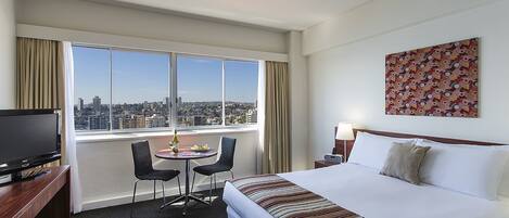 A hotel room with a large window, a bed, a desk with a TV, a chair, and a table with a bottle and fruit.