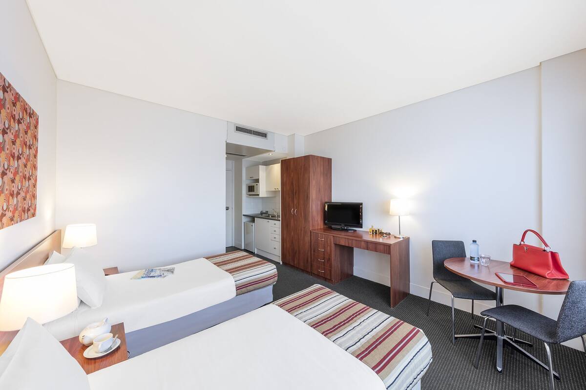 Habitación de hotel con una cama, un escritorio con televisor, una silla, una mesa con un bolso rojo y una kitchenette al fondo.