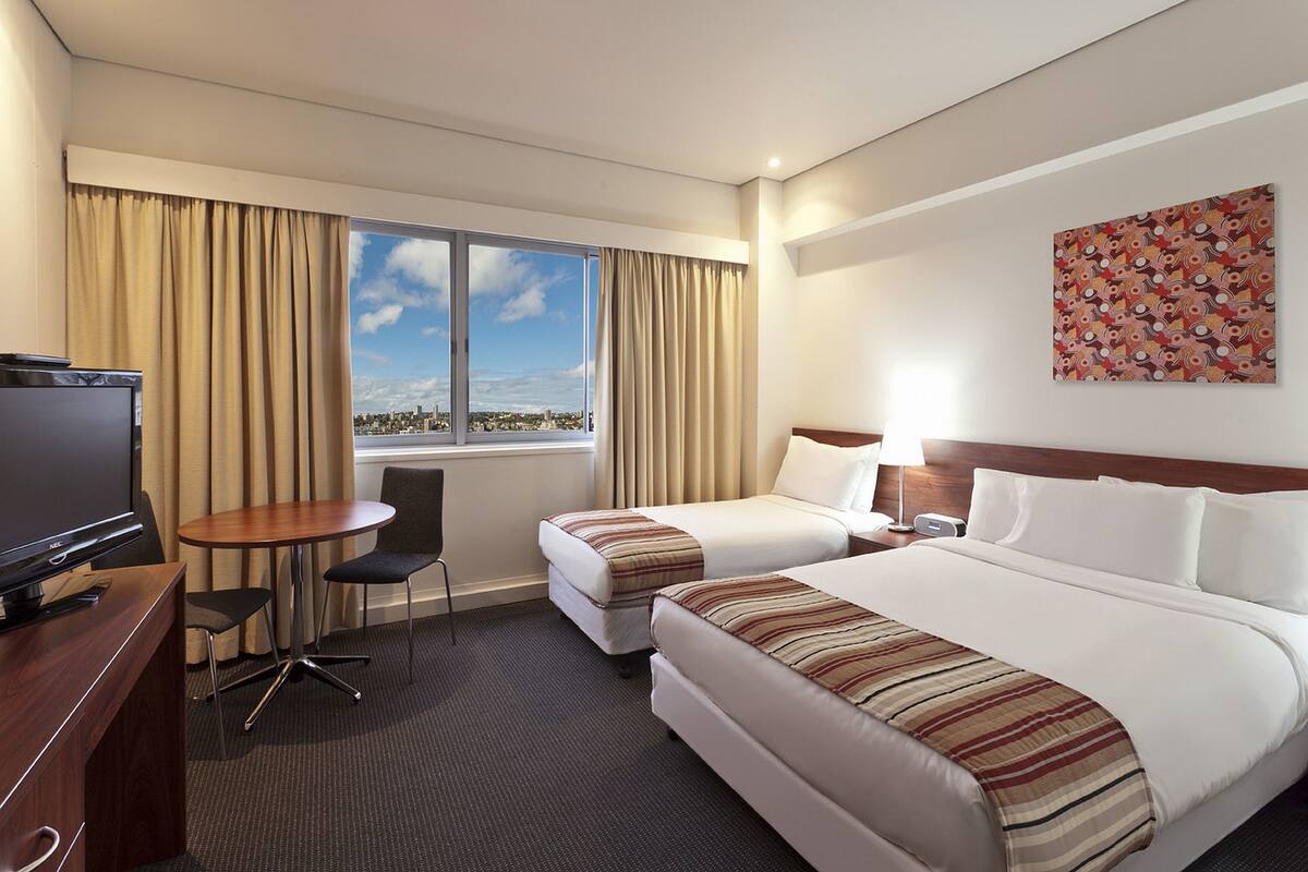 Habitación de hotel con dos camas, un escritorio, una silla, un televisor y una ventana con vista a la ciudad.