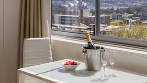 Une table avec une bouteille de champagne, deux verres et un bol de fruits rouges. On aperçoit à travers la fenêtre un paysage urbain avec un pont et un opéra.