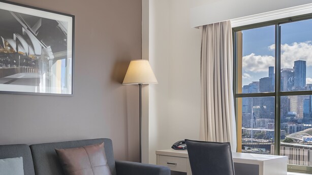 A modern hotel room with a grey sofa, a desk, and a chair. There is a large window with a city view. - Oaks Sydney Goldsbrough Suites (Pyrmont)