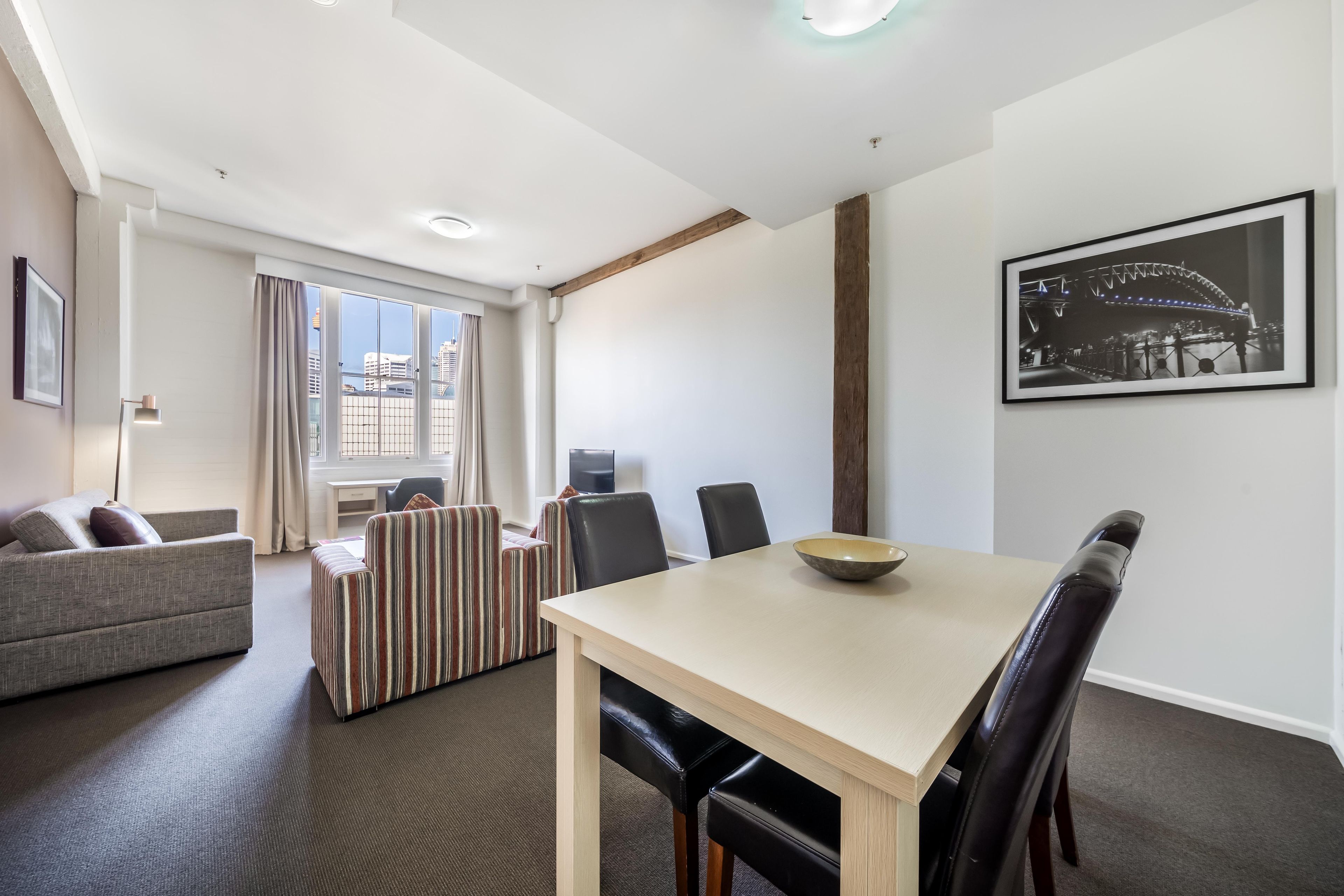 A modern hotel room with a dining area, a sofa, a TV, and a cityscape view.