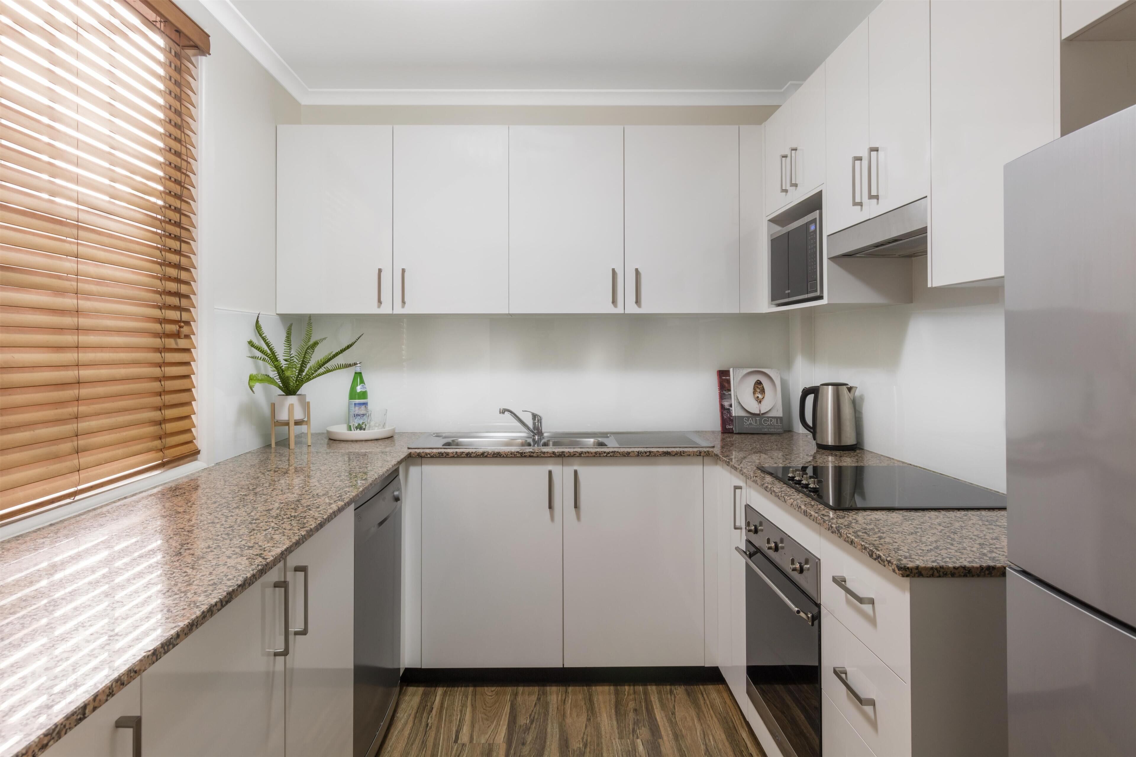 Una cocina moderna con gabinetes blancos, encimera de granito, microondas integrado y refrigerador de acero inoxidable.