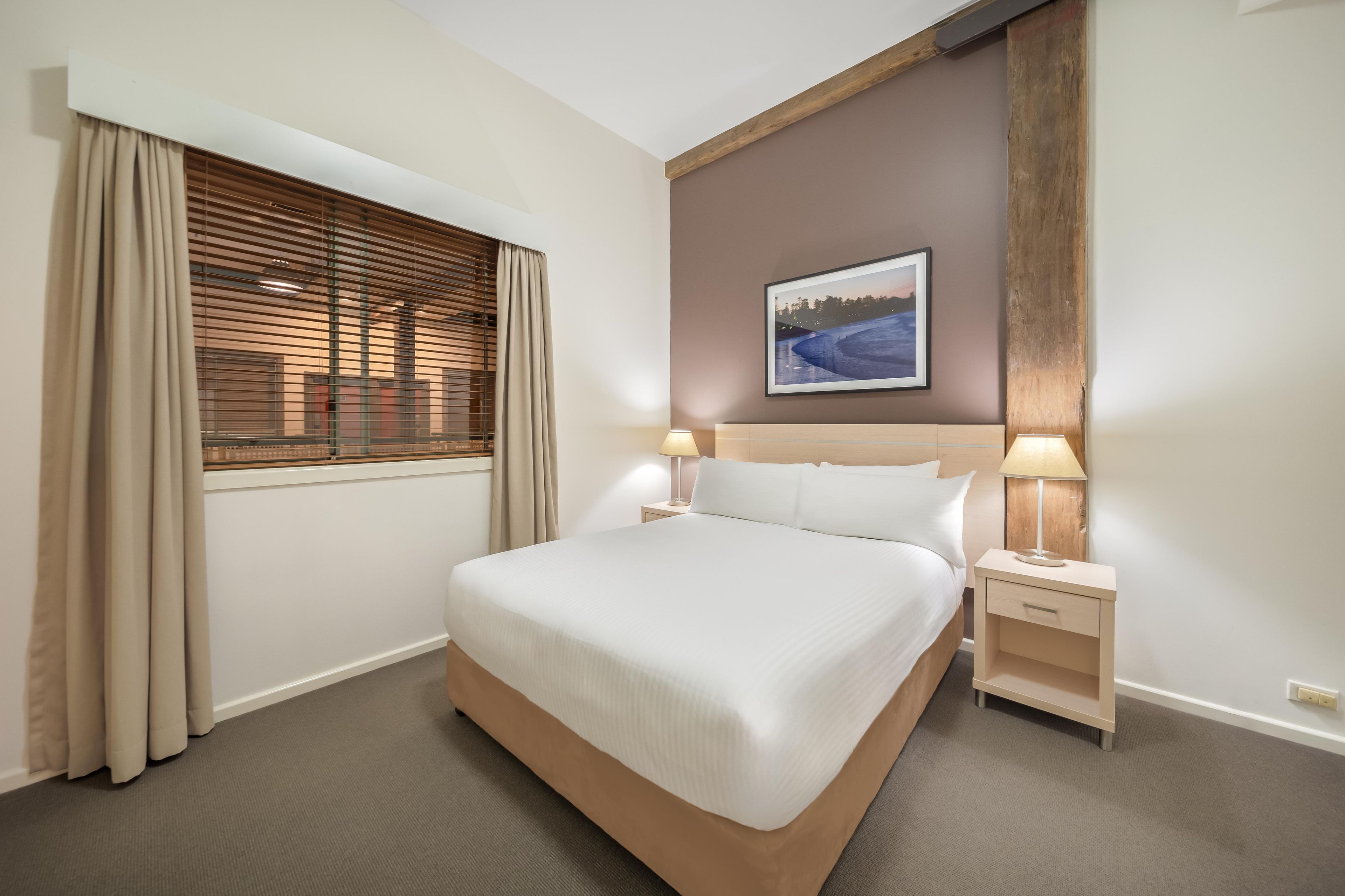 Habitación de hotel con una cama grande, dos mesitas de noche con lámparas, una ventana con persianas y un cuadro en la pared.