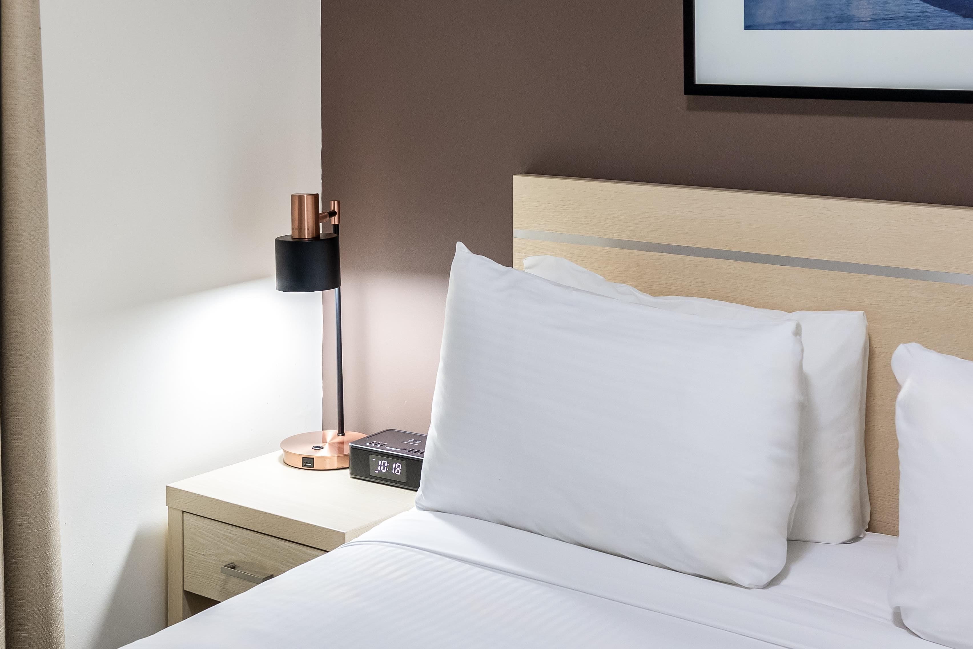 Ein ordentlich bezogenes Bett mit weißen Leinen und Kissen, ein Nachttisch mit Lampe und digitaler Uhr sowie ein gerahmtes Bild an der Wand.