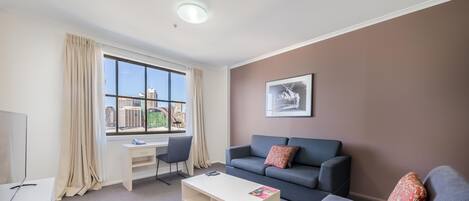 A modern living room with a sofa, a coffee table, a desk, and a large window offering a city view.
