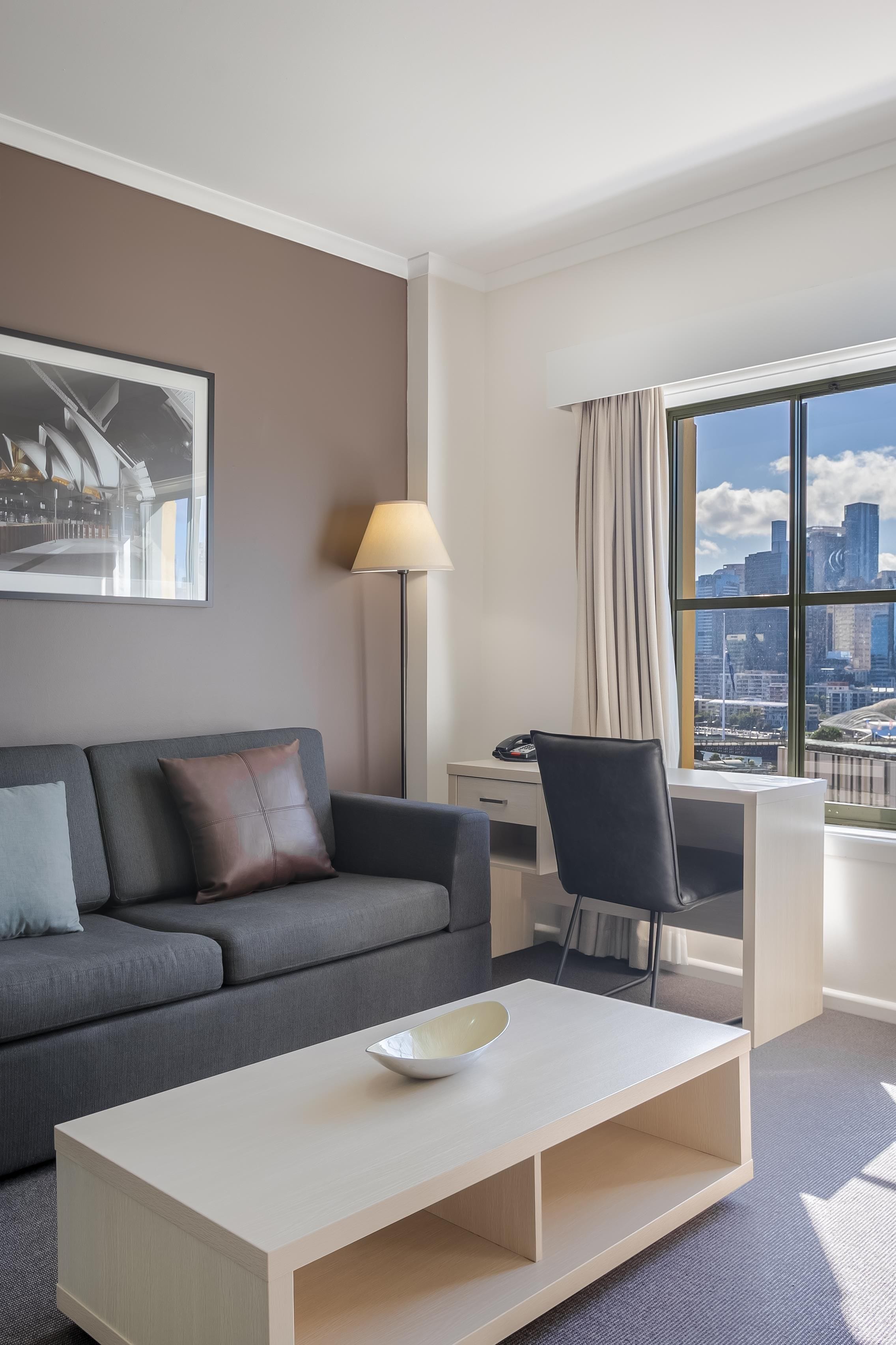 Et moderne hotelværelse med en grå sofa, et skrivebord, en stol, en lampe og et stort vindue med udsigt over byen.