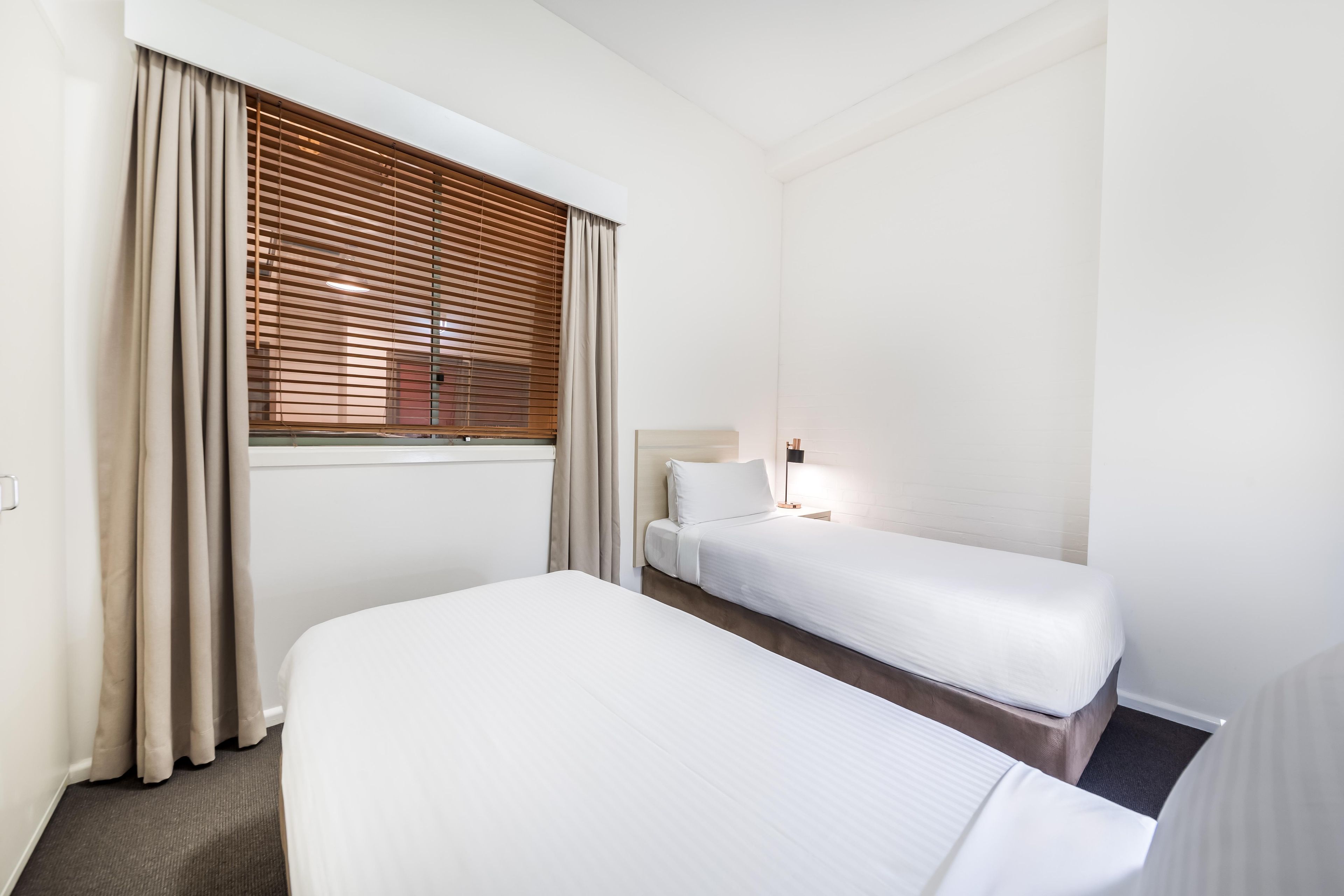 A hotel room with two single beds, a window with blinds, a bedside lamp, and a carpeted floor.