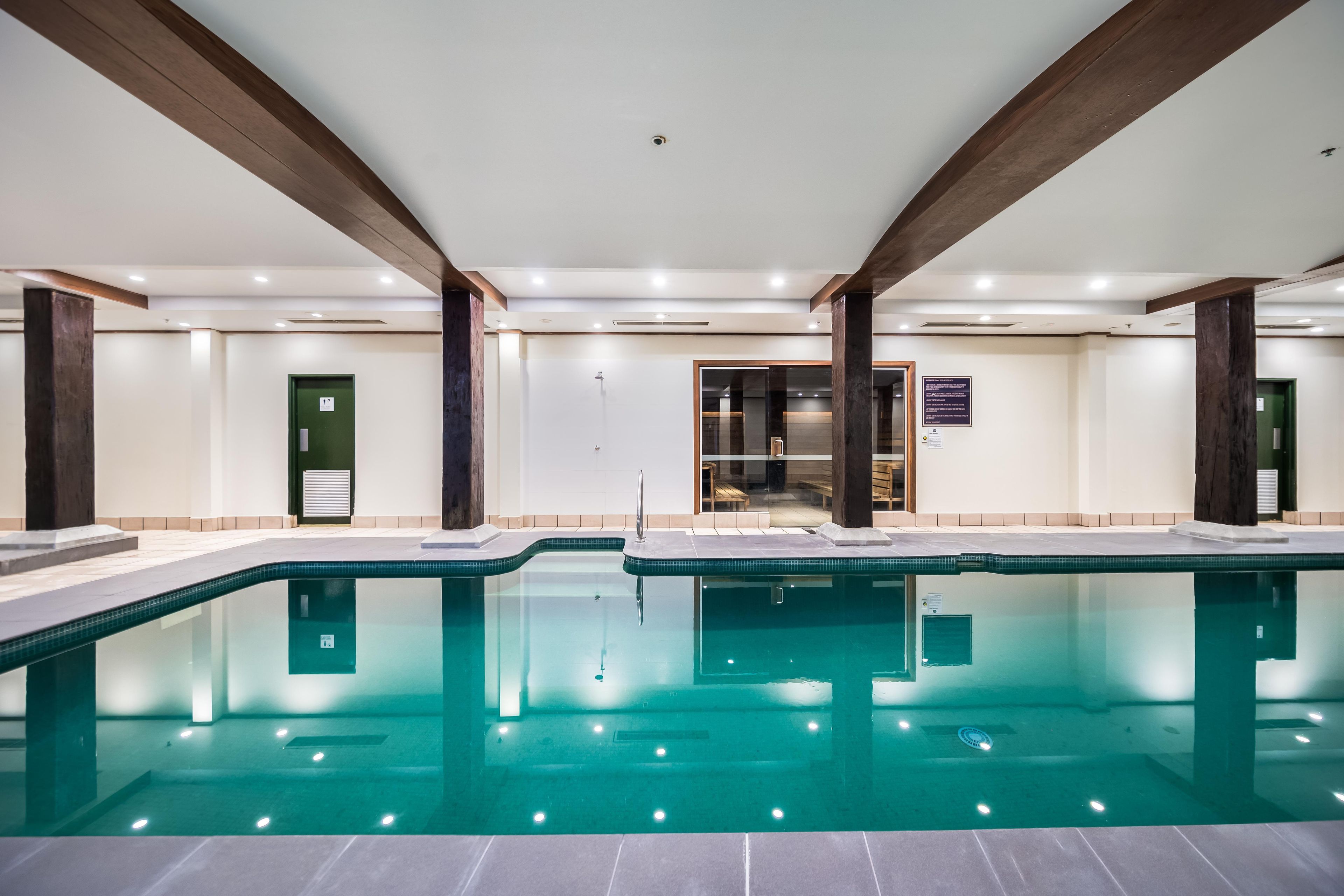 Piscina con solárium de baldosas, vigas de madera y cerramiento de vidrio para vestuarios.