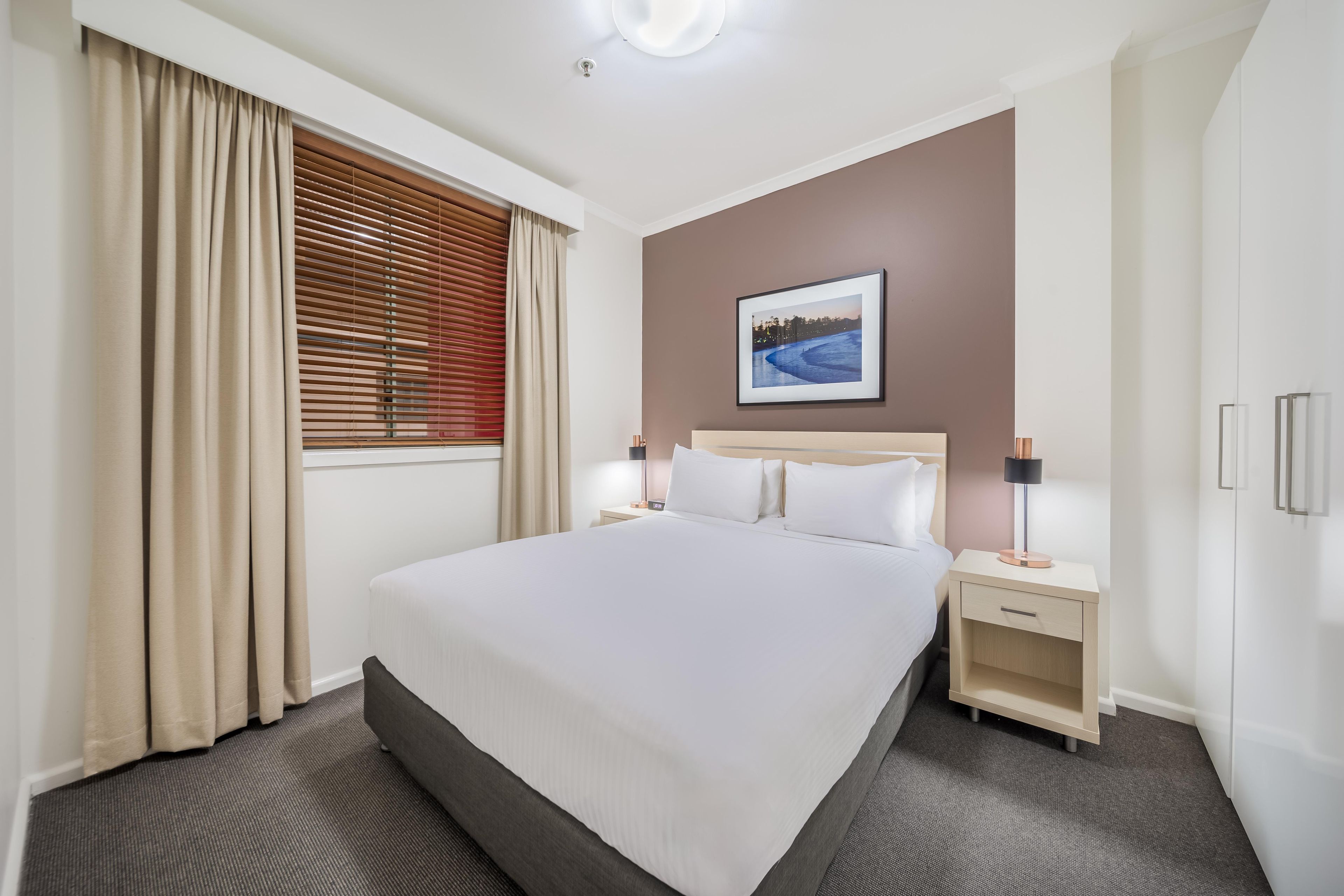 Une chambre d’hôtel avec un grand lit, une table de chevet, une lampe, un tableau au mur et une fenêtre avec des rideaux.